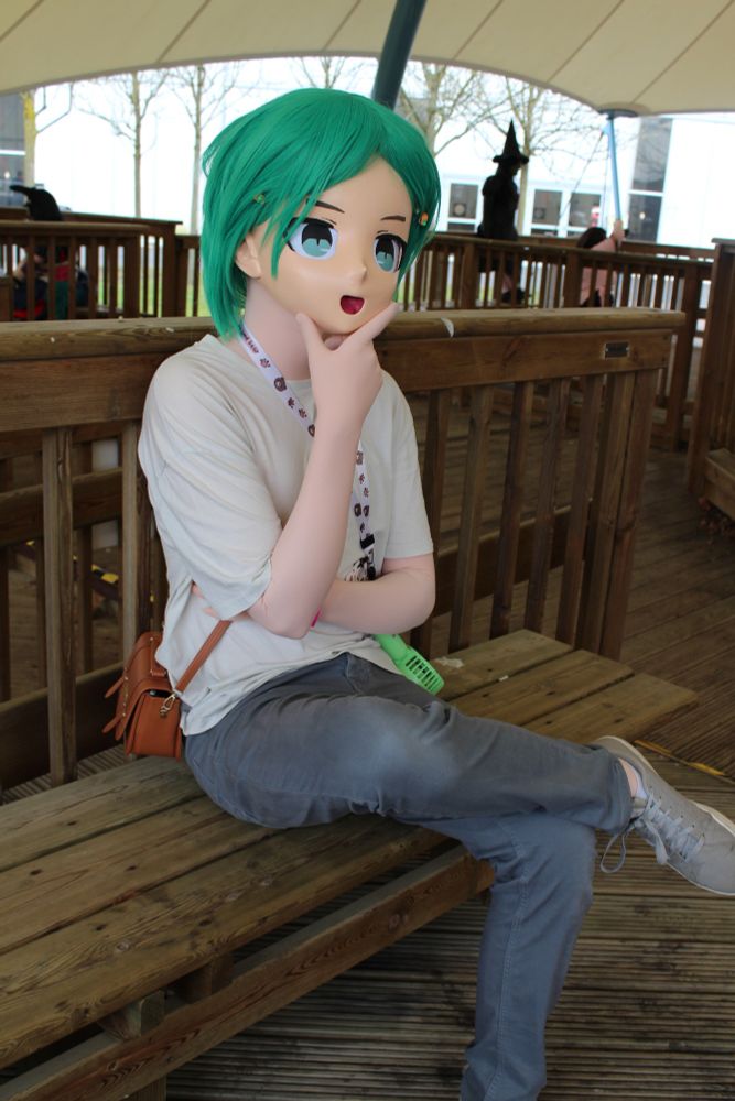 A photo of a green haired kigurumi character sat on a wooden bench underneath a large tent. They have their hand on their chin deep in thought. 