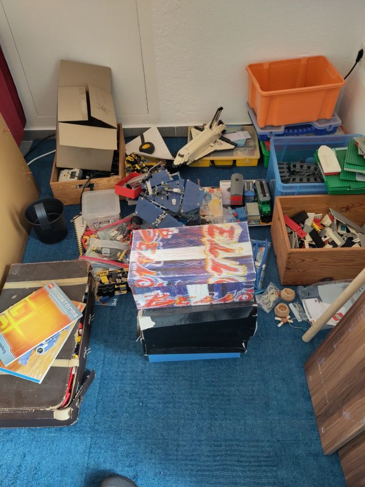 Ecke von einem Raum mit einem Teppich auf der Kisten voll Lego sowie ein Marsrover und das Spaceshuttle aus Lego liegen 