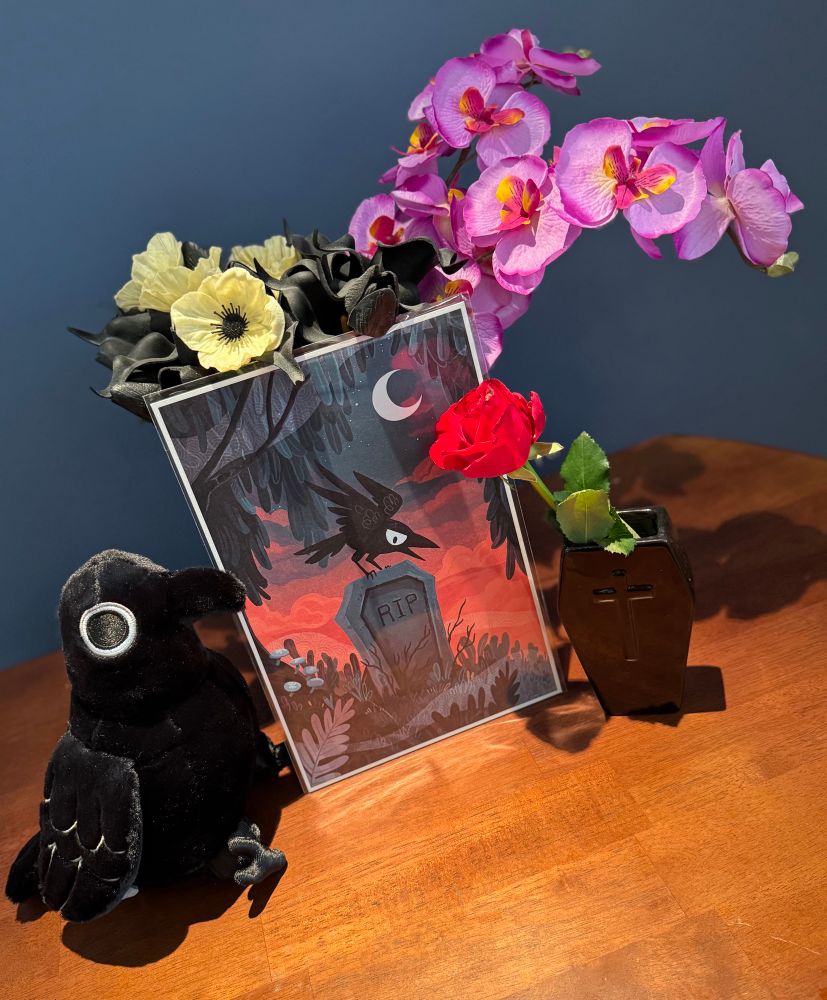 A vase of fake flowers behind an art print of a crow on a tombstone, a crow plushy, and a real red rose in a coffin-shaped vase. 
