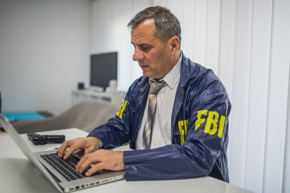 A Caucasian man in a navy blue jacket with yellow "FBI" lettering is seen typing on a computer.