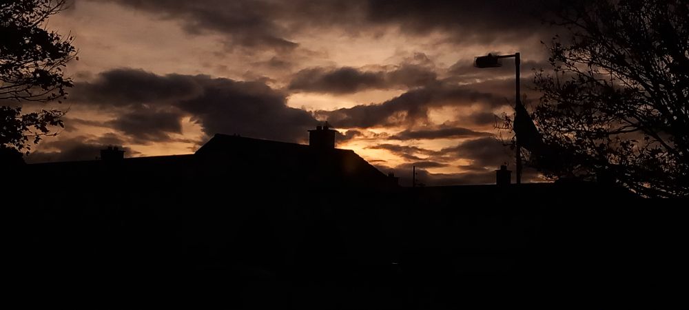 A photo of clouds. The bottom half of the frame is covered by the silhouette of houses. The top half is filled with many clouds; Big lumps, small streaks, dots & dashes. The sky behind the clouds is a vibrant display of bright oranges & yellows towards the center of the frame, fading to a dull grey further outward.