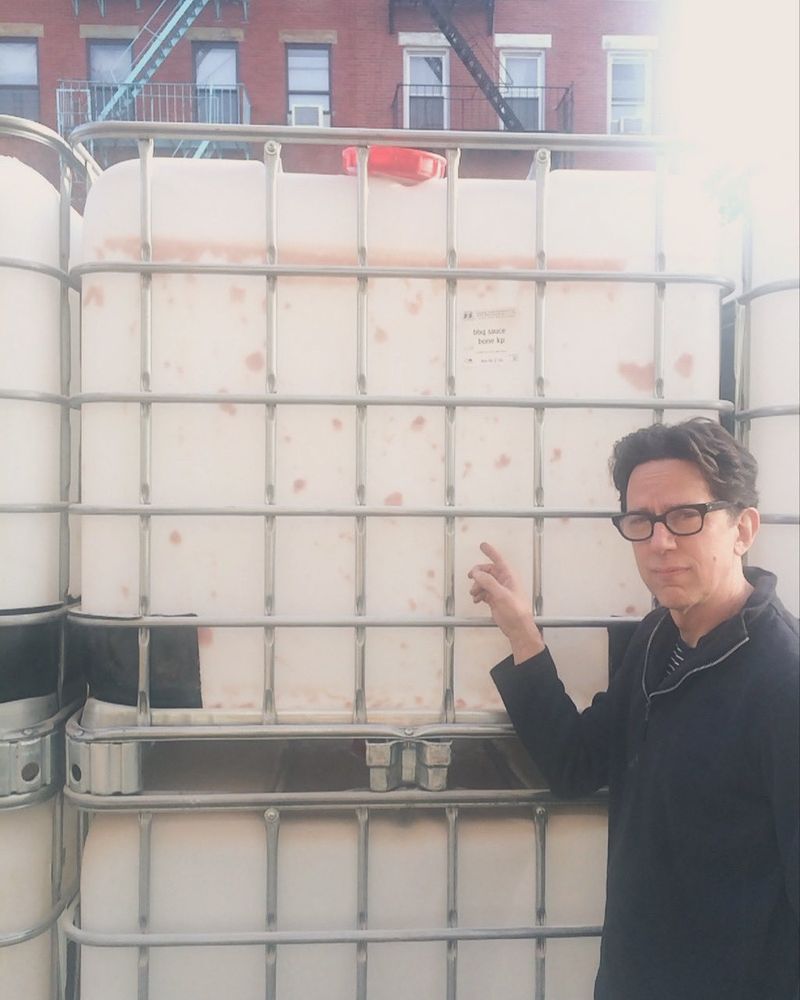 John Linnel of They Might Be Giants pointing at a white IBC container. The photo is overexposed.