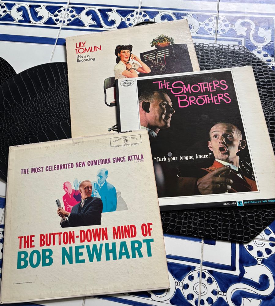 Three records one by Lily Tomlin called “This is a recording”, one by the Smothers Brothers called “Curb your tongue, knave,” and “The button down mind of Bob Newhart” on a white and black table with black placemats.