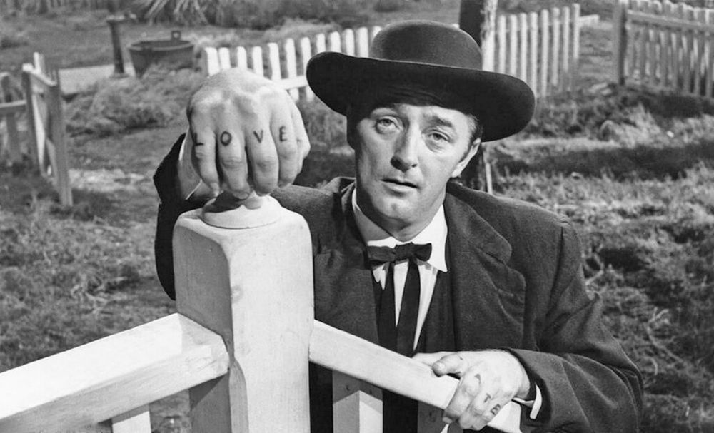 A still from Charles Laughton only film, Night of the Hunter. Mitchum, in black hat and string tie grasps the finial of a porch bannister. He reveals the tattoos on his fingers, "Love" and "Hate."