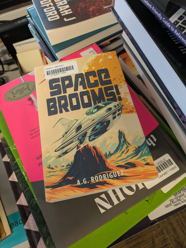 A stack of books of all shapes, sizes, and colors, with a paperback copy of SPACE BROOMS! by A.G. Rodriguez sitting prominently on top. The books cover is a mix of yellows, browns, and oranges with a big, retro futuristic spaceship on the cover. 