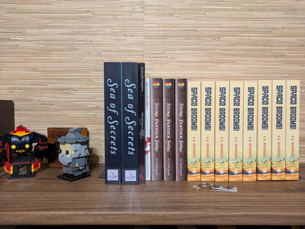 A pic of all my books, or books I've been featured in, sitting on top of a brown bookshelf. Next to them are two Lego figurines of Gandalf and the Balrog from Lord of the Rings. 