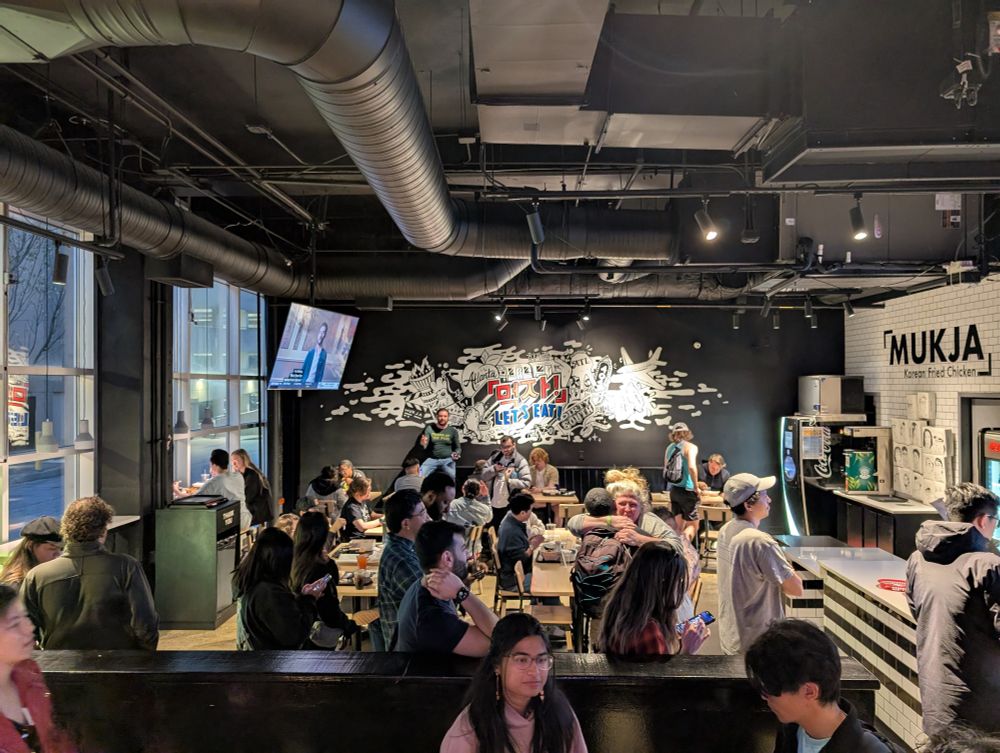 A group of people sitting at tables in a restaurant. Several people are waiting in line