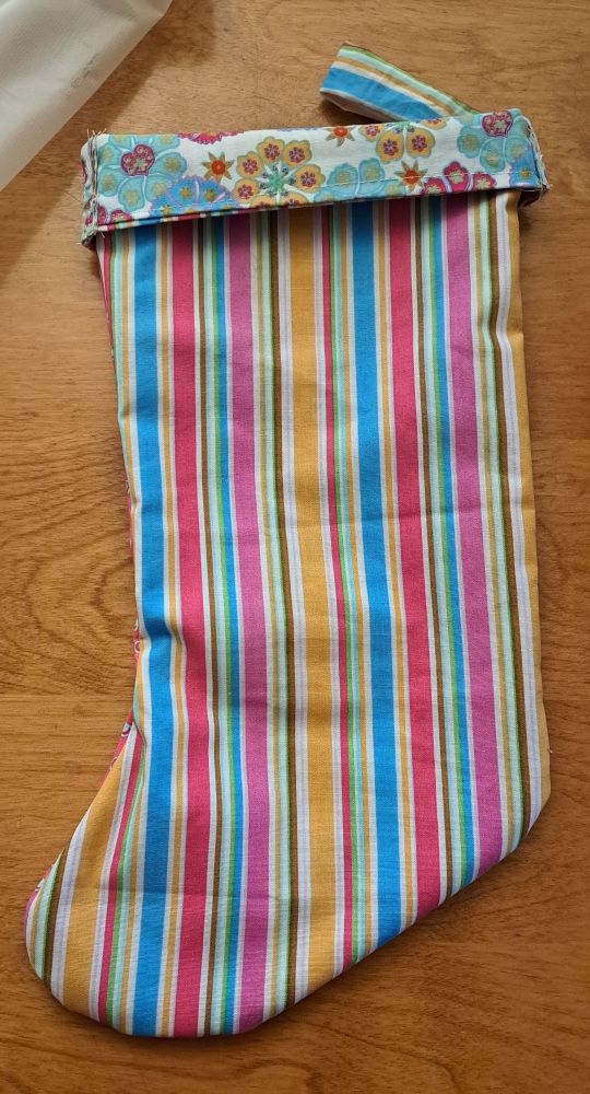 One side of a Christmas stocking. The pattern on this side is multicoloured stripes of varying thickness, and this pattern repeats on the little hanger piece. There is a colourful floral pattern as the cuff. 