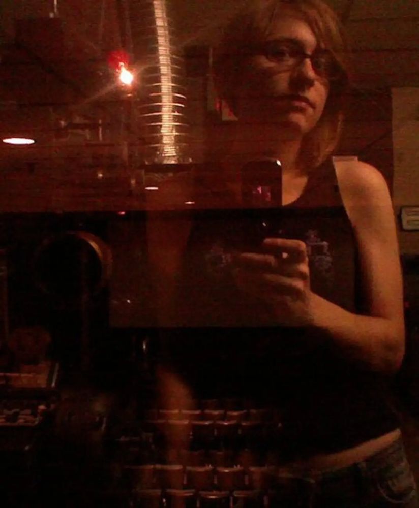 Mél in 2009, wearing a black tank top and jeans, is taking a selfie of themselves reflected in the projection booth window of a cinema, where they worked at the time. Through their reflection you can see the seats and a bit of the screen.