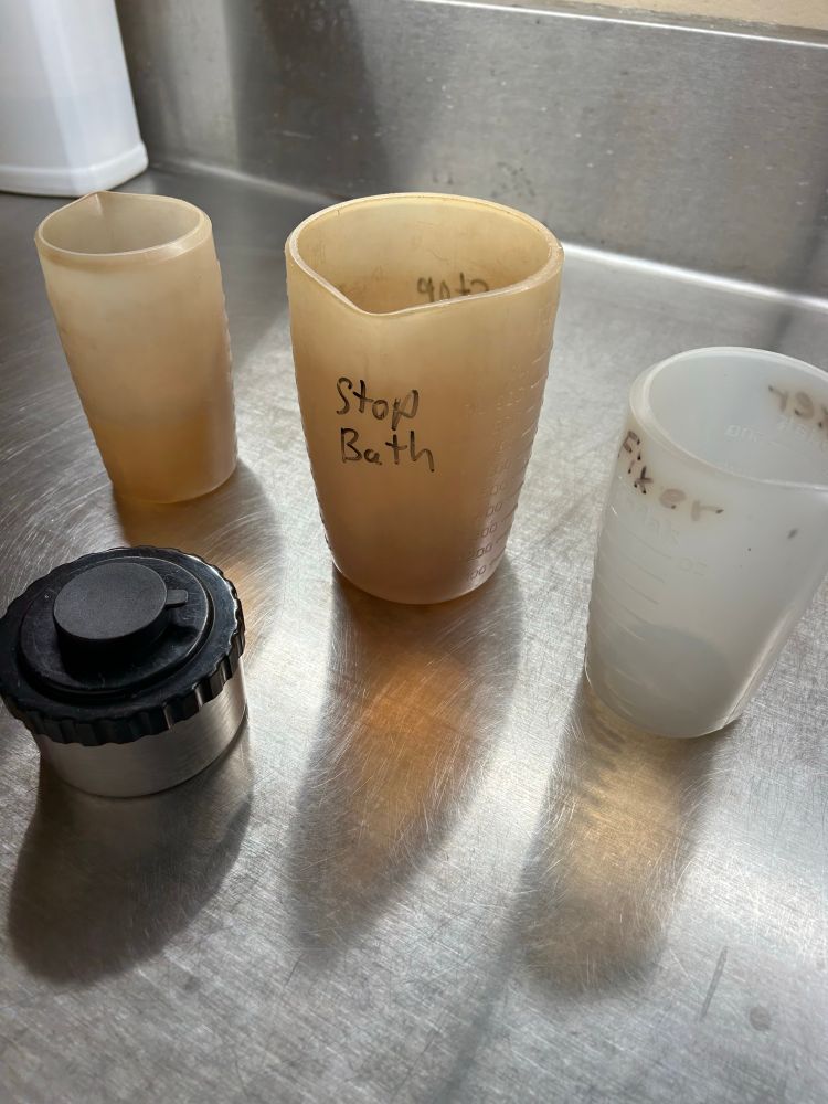 A light-tight canister sitting in a stainless steel sink holding a roll of film. Behind it are three measuring cylinders of developer, stop bath, and fixer.
