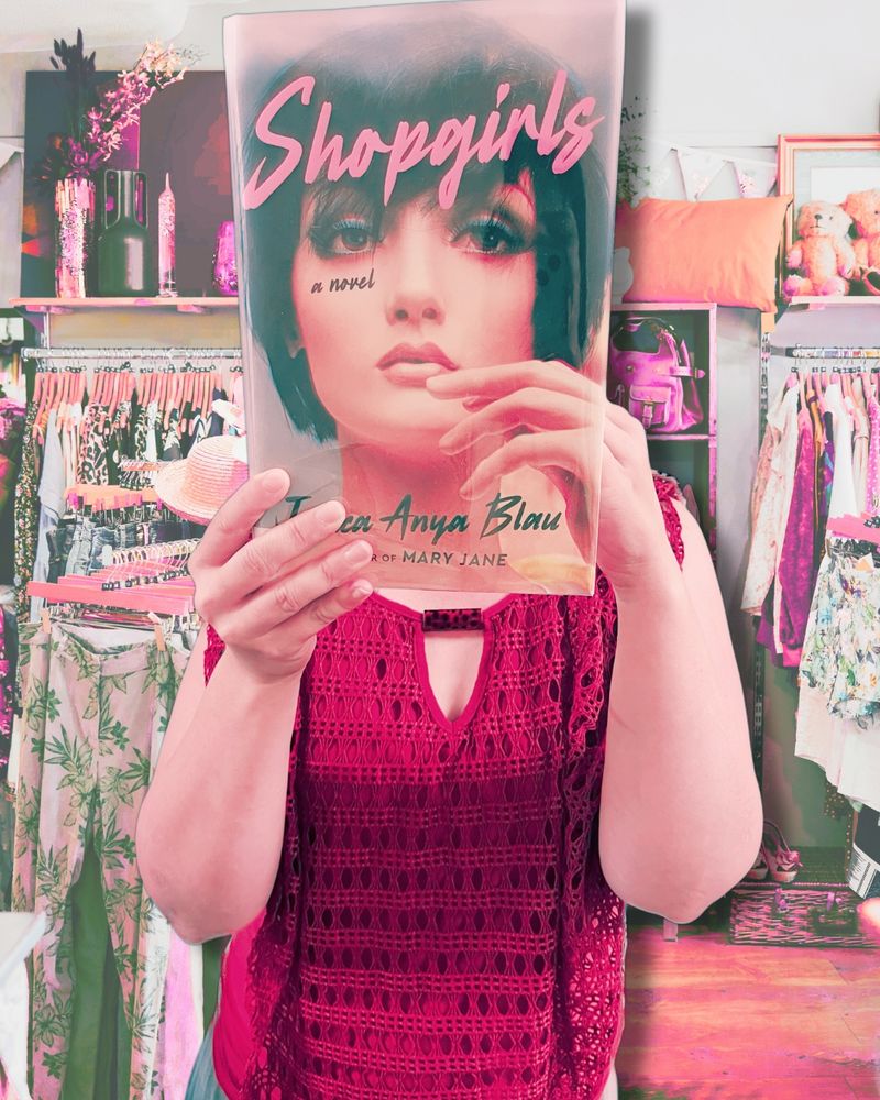 Bookface photo of staff member with the book “Shopgirls” by Jessica Anya Blau. Bookface photos are when staff pose with a book cover to align themselves visually with the image on the cover to create an optical illusion. 