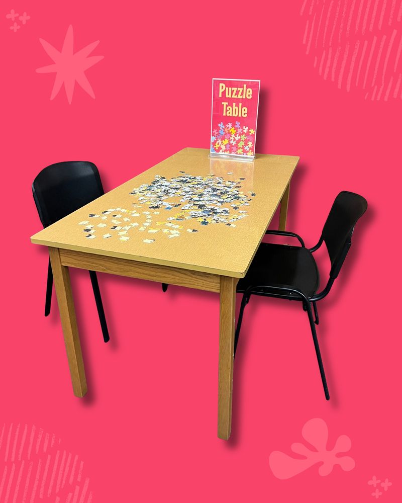 Photo of a wooden table and two chairs. The table has puzzle pieces scattered on top and a sign that reads Puzzle Table. Image has a bright pink background with wonky shapes.