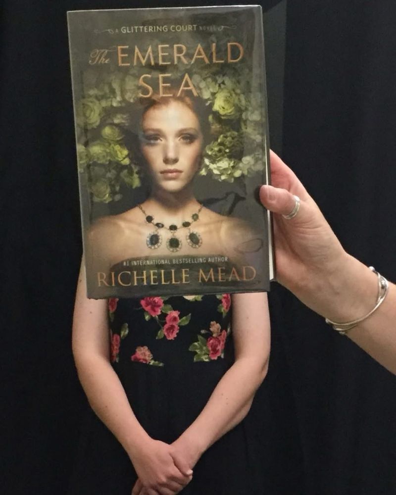 Photo of a woman with the book “The Emerald Sea” by Richelle Mead.Bookface photos are when staff pose with a book cover to align themselves visually with the image on the cover to create an optical illusion. 