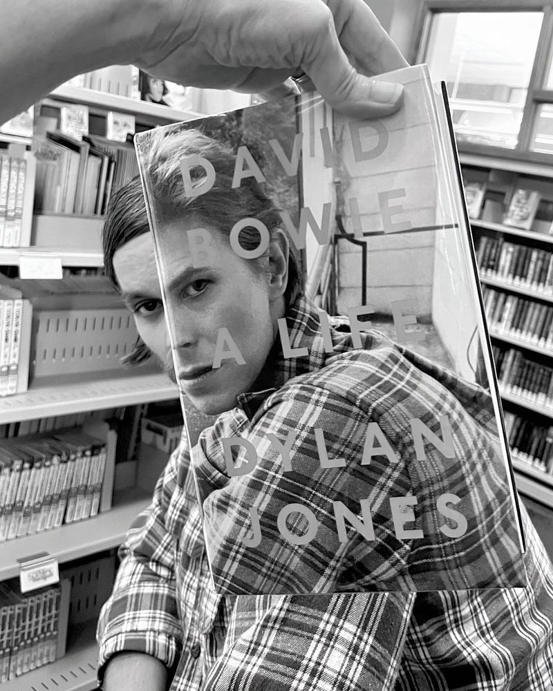 Bookface photo of a staff member with the book “David Bowie: A Life” by Dylan Jones. Bookface photos are when staff pose with a book cover to align themselves visually with the image on the cover to create an optical illusion. 