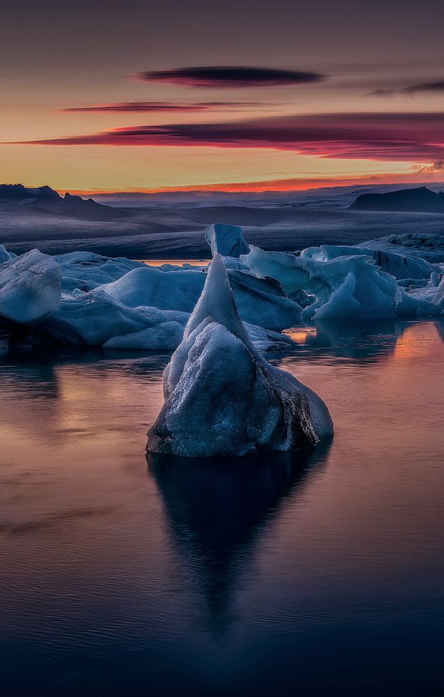 Sunset scene showing floating icebergs at the Glacier Lagoon.  The iceberg in foreground have pointy shape, with dark reflection in the water. In background we see the glacier layers and the red clouds. 