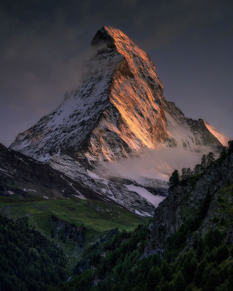 The Matterhorn summit throning over the Alps, illuminated by orange/red sunset glow. The mountain looks very imposing. In foreground there’s more mountain layers covered by trees and vegetation. The mountain is also covered by thin layer of snow. The sky is quite neutral, with soft clouds passing over the summit. 