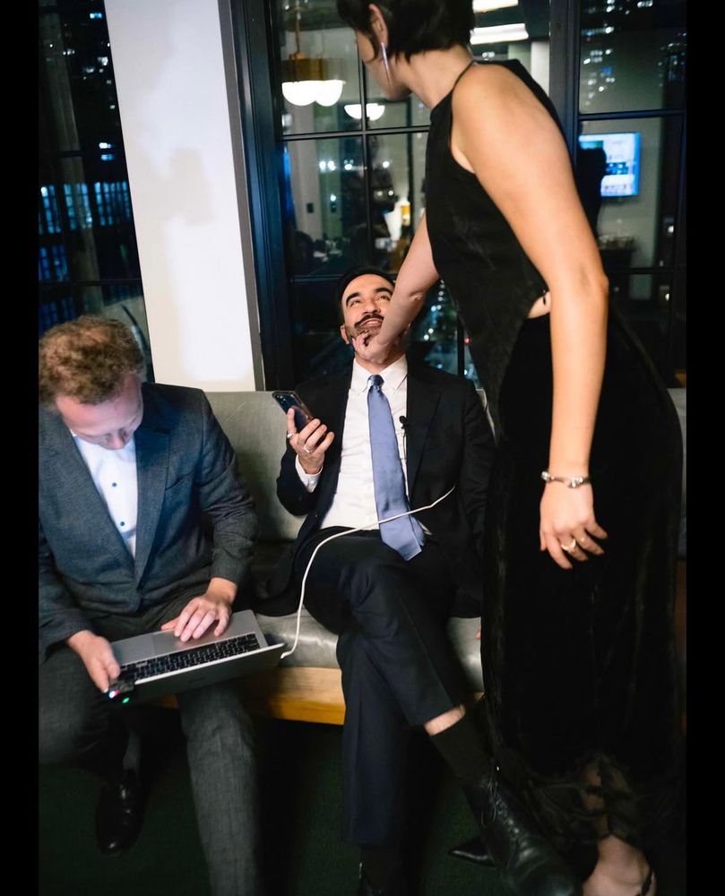 New York mayor-elect Mamdani seated with his wife standing over him, holding his chin as he smiles up at her, happy as a lamb 