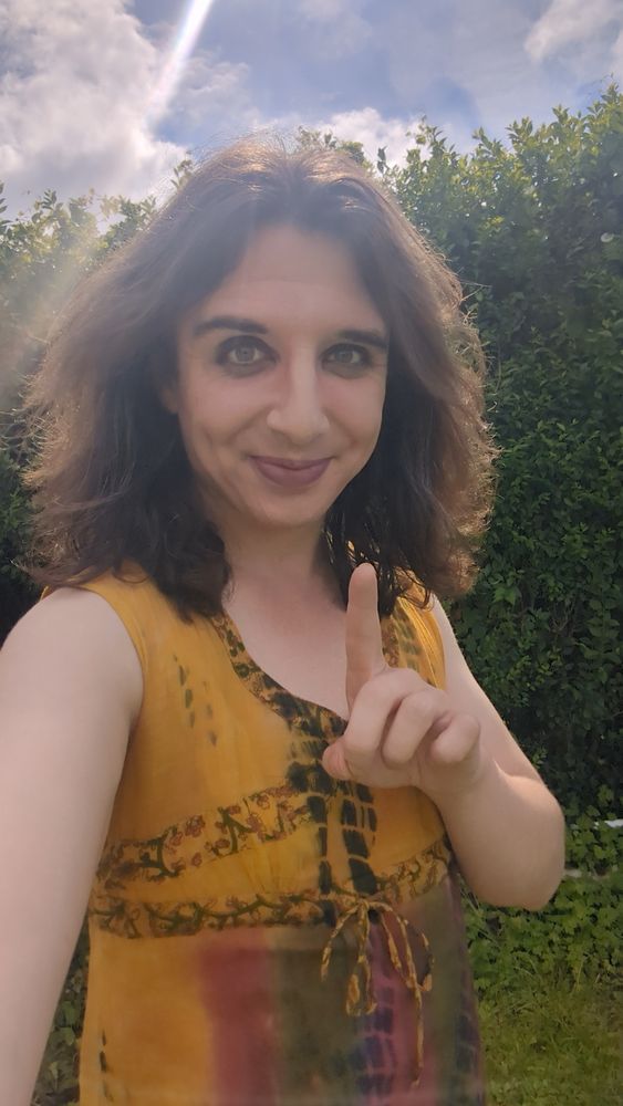 Neilly in the garden with the sunshine creating a lens flare behind her, in a yellow sundress with a single index finger raised 