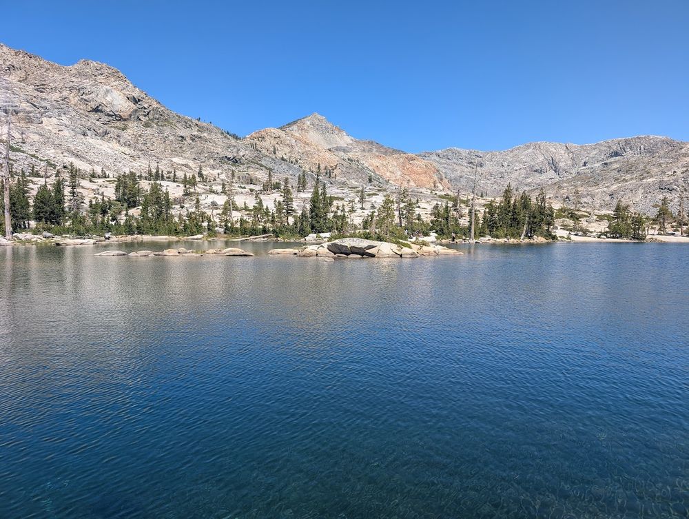 Photo of parts of Twin Lakes along the hike to Island Lake near Wrights Lake. There's a stone "island" in the middle. Water is very clear and blue. Trees on the shore. Mountains in the background.