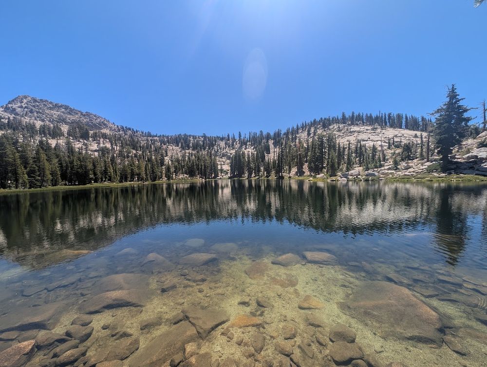 Photo of Grouse Lake up at 8k feed, very clear water, so you can see rocks beneath the surface. Lake surface is clear and you can see the reflection of the mountains and trees on the other side of the lake.