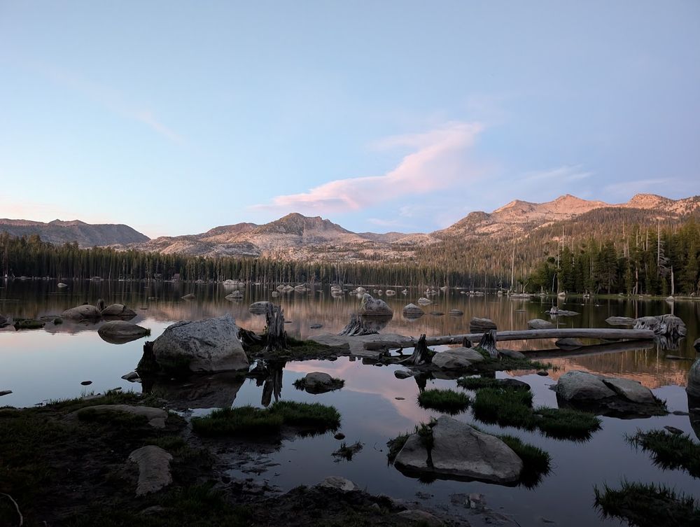 Another photo of the clear reflective lake, at dusk, with mountains in the background, and many boulders sticking up throughout the lake.