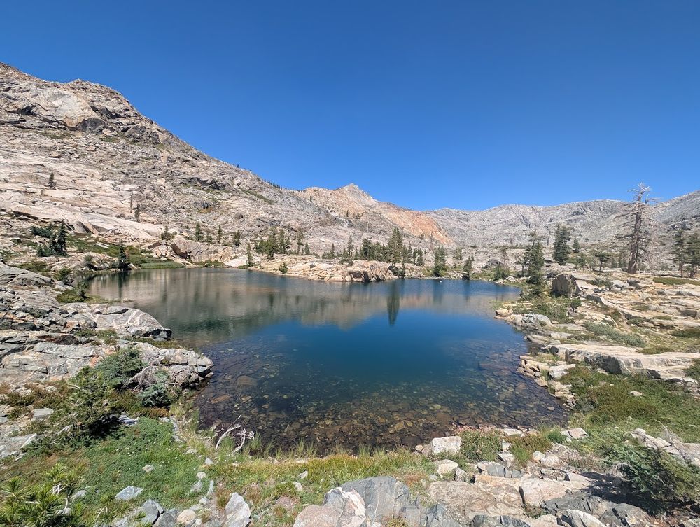 Wide angle photo of Boomerang Lake, a small mountain lake vaguely shaped like a boomerang. Or Mickey Mouse. Mountains in the background. Some trees dotted around the shore