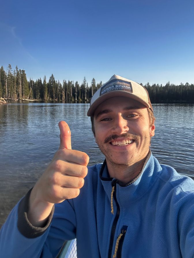 Photo of a guy in a kayak or canoe smiling and giving a thumbs up. He has a mustache, and a blue zip fleece, and is wearing a Sierra Nevada hat.