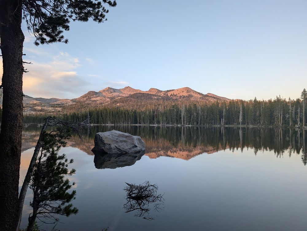 Photo of a very clear, reflective lake with a giant bolder in the middle, a tree is in the foreground on the left, and in the distance are mountains that are then reflected upside down in the lake.