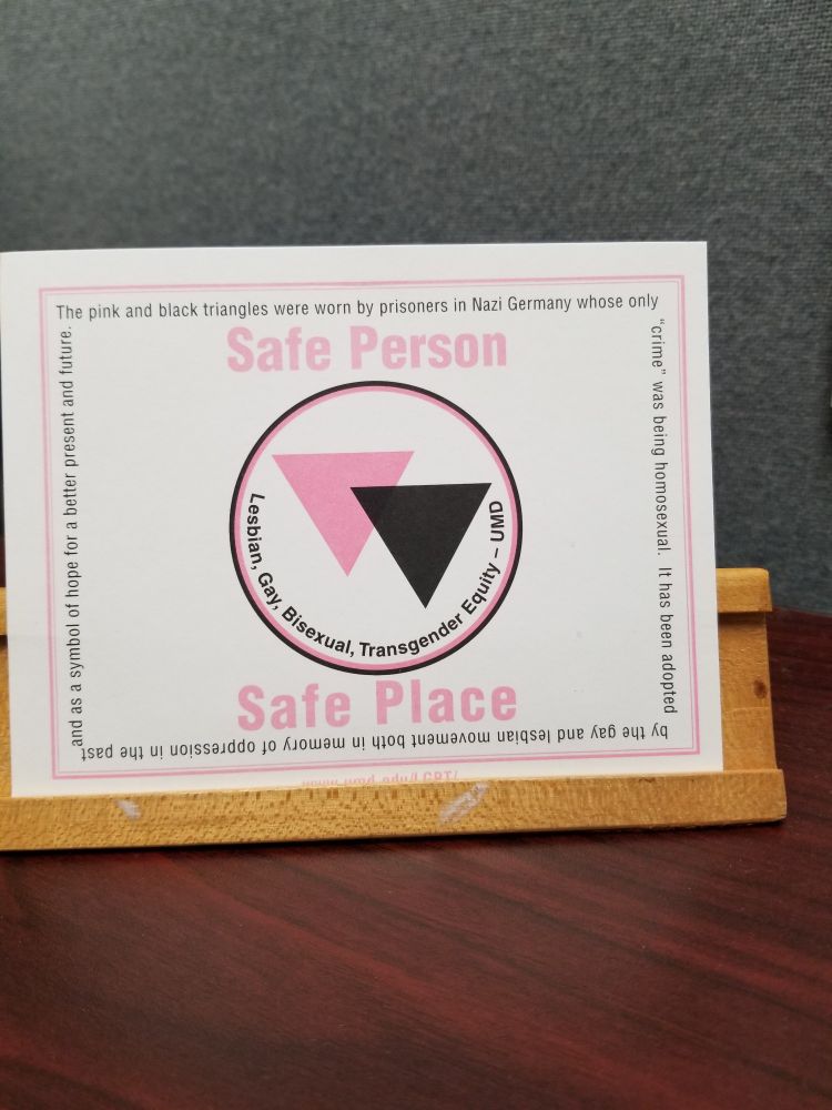 Safe Person Safe Space postcard from the Office of Lesbian, Gay, Bisexual and Transgender Equity, UMD, with an image of black and pink triangles in a circle. Text reads: "The pink and black triangles were worn by prisoners in Nazi Germany  whose only 'crime' was being homosexual.  It has been adopted by the gay and lesbian movement in memory of oppression in the past and as a symbol of hope for a better future. "