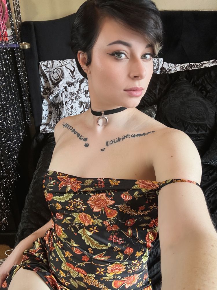 Woman with short black hair wearing a black spaghetti strap dress with an orange floral pattern and a moon choker necklace, trying to make a somewhat attractive facial expression 😅