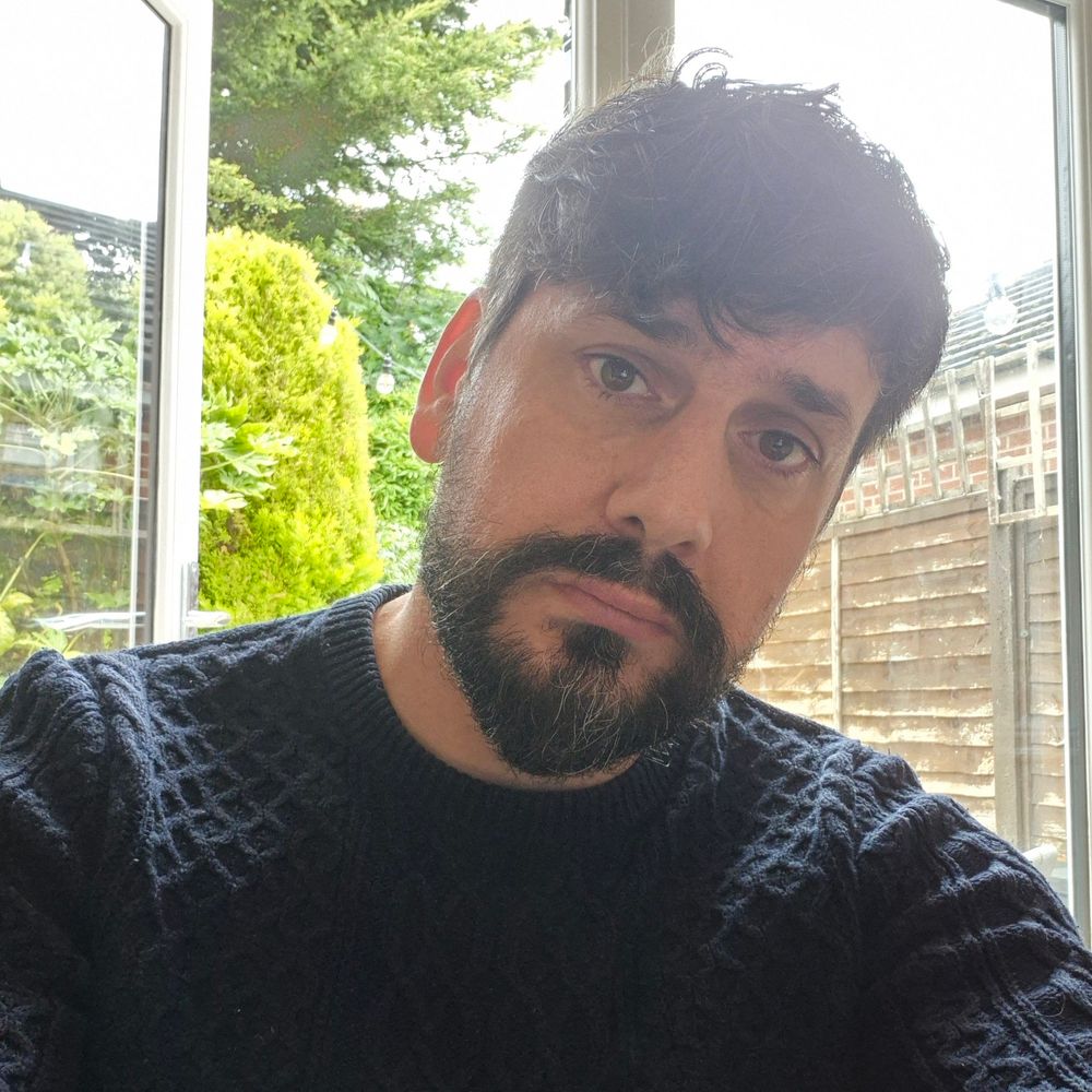 A Caucasian man with black hair and beard sits looking at the camera wearing a navy woollen sweater. He has a thoughtful expression.