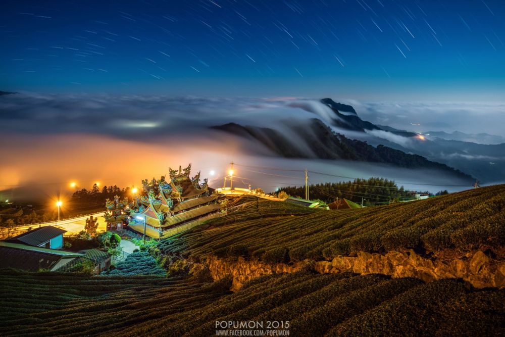 Das Foto zeigt eine atemberaubende Nachtaufnahme einer Gebirgslandschaft in Taiwan. Im Vordergrund sind die geschwungenen Reihen einer Teeplantage zu sehen, die sich über die Hänge ziehen. In der Mitte des Bildes steht ein prachtvoll beleuchteter, traditioneller chinesischer Tempel mit kunstvoll verzierten Dächern, die in leuchtenden Farben schimmern. Nebel strömt wie ein weiches Meer durch die Täler und hüllt die umliegenden Berge in eine mystische Atmosphäre. Am Himmel sind feine, kreisförmige Lichtspuren der Sterne zu erkennen – ein Zeichen einer Langzeitbelichtung. Die gesamte Szene wirkt friedlich, magisch und fast unwirklich schön.
