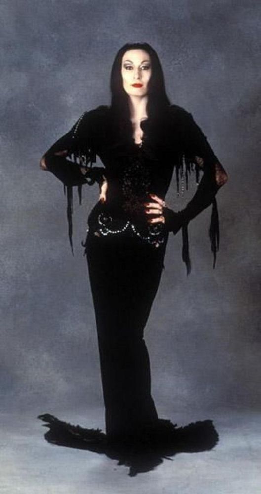 a full length shot of Anjelica Huston as Morticia Addams, wearing a long, slim-fitting black dress woth sequins around the hips and bits of lace and netting dripping from her sleeves