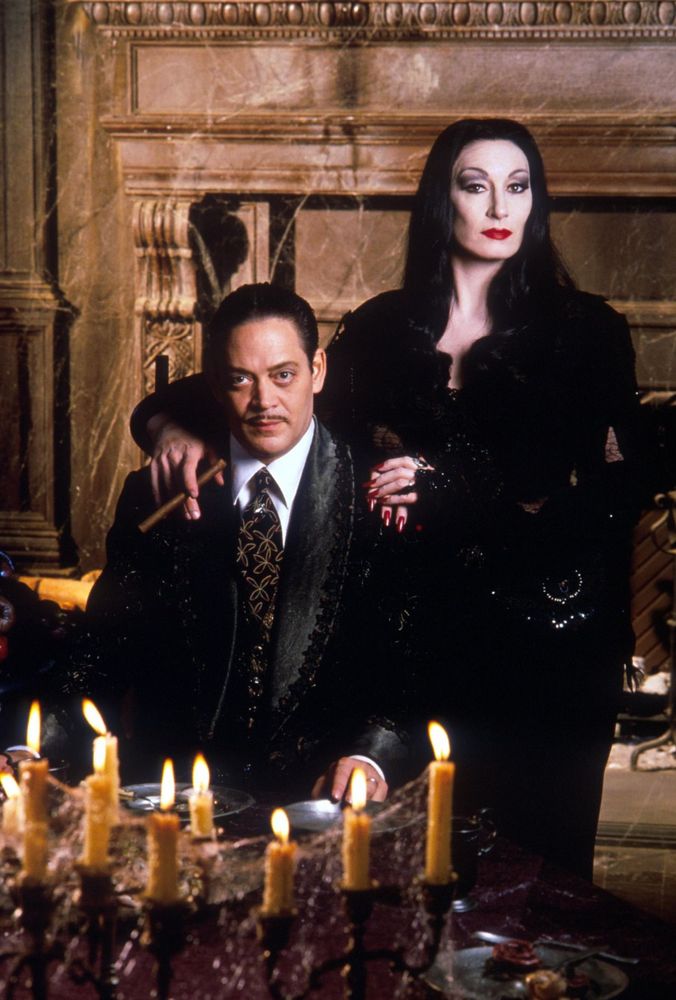 A portrait shot of Gomez and Morticia Addams; he is seated at a table, she is standing behind him with her hands on his shoulder