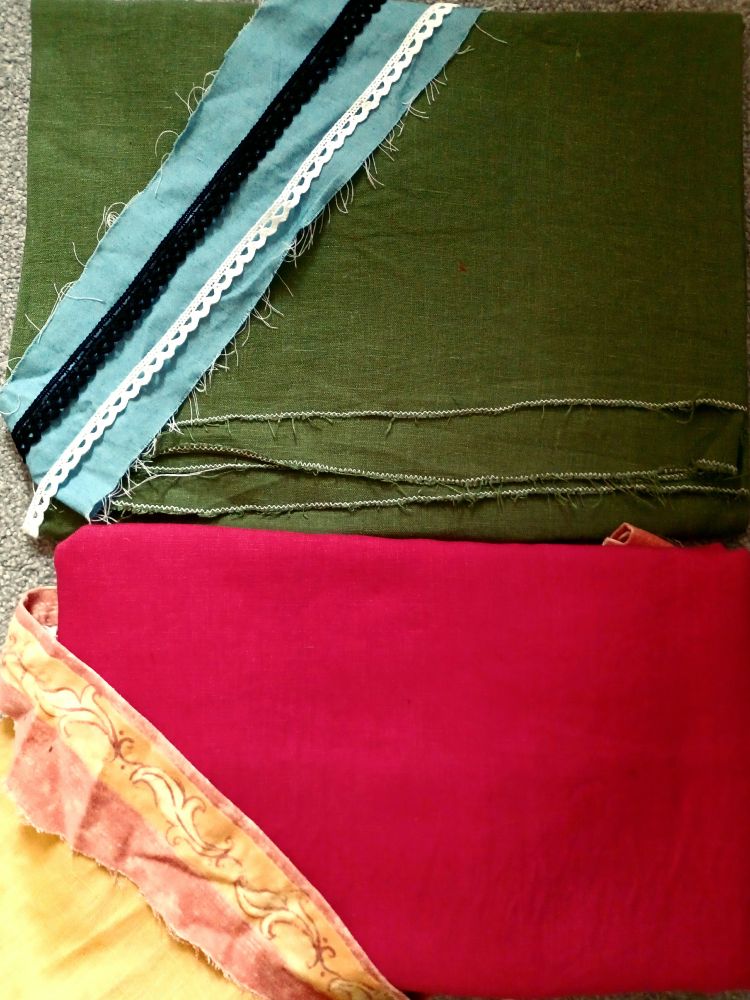 a folded piece of green linen with a scrap of blue cotton and some black and white lace on it, and next to it a folded piece of red linen with a pattern orange scrap and some yellow linen on it