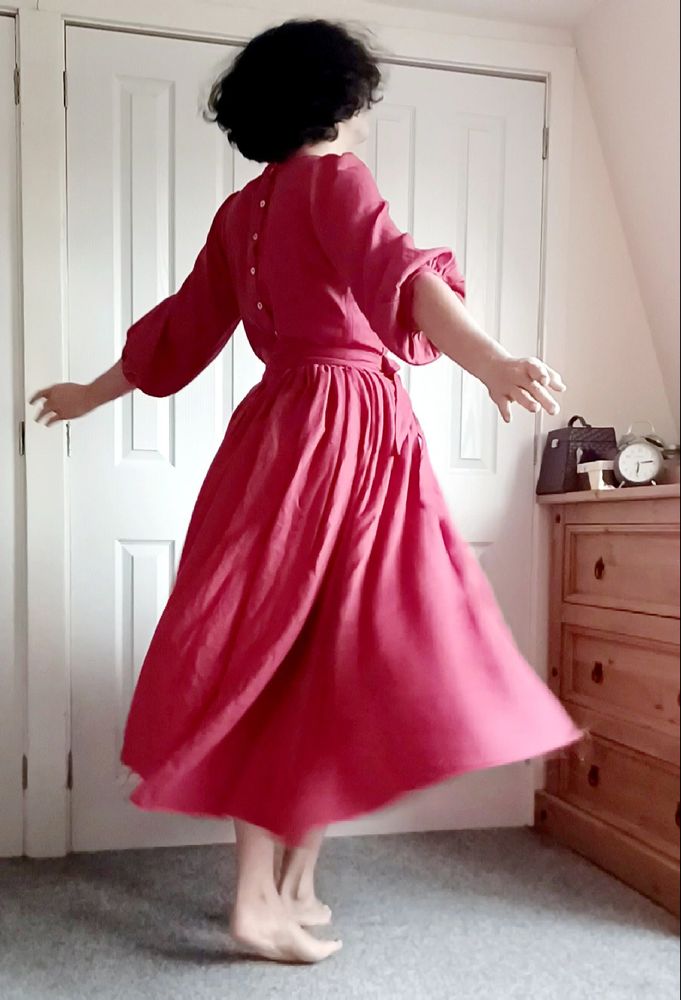 side back view of a white person with short, fluffy dark hair wearing a calf-length red dress with a very swishy skirt, a fitted waist, and puffy elbow-length sleeves
