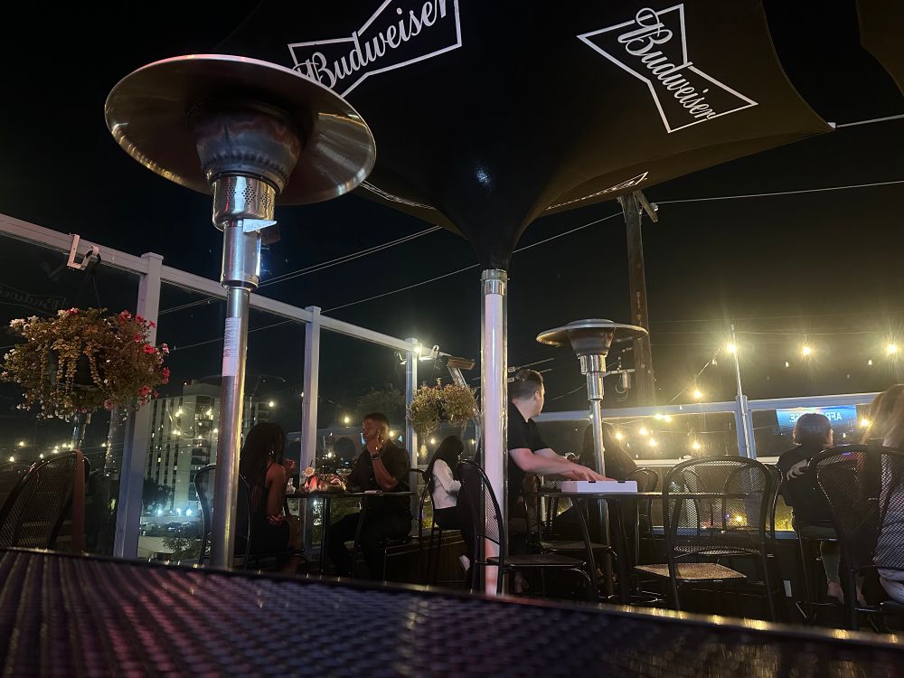 A view of the rooftop patio with tall tables and heaters. Glowing patio lights, baskets of flowers and people having an amazing evening. 