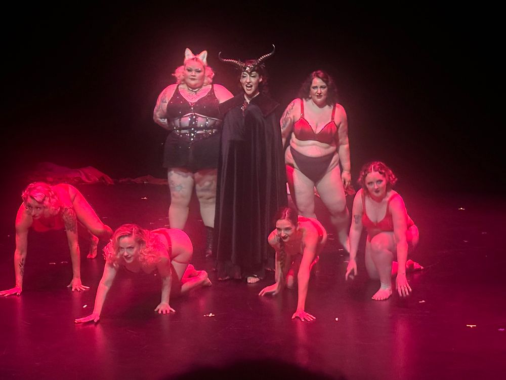 Five ladies in red undercrackers mid floor work around a black cloaked devil, a cat in black undercrackers at her side. 