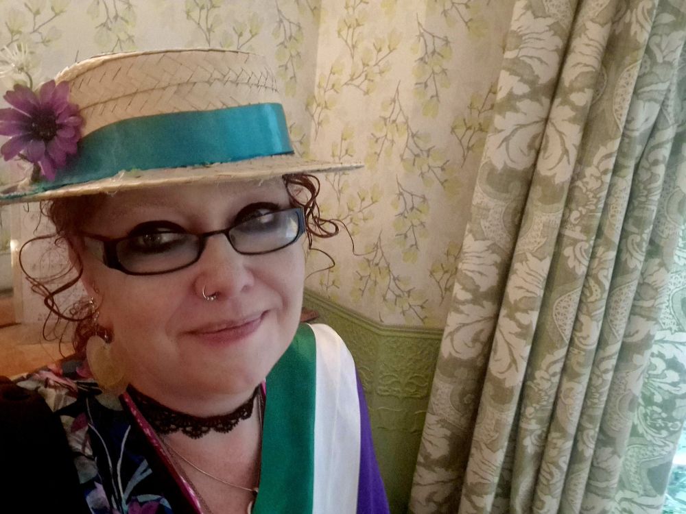 A picture of me in the historic parlour at the Pankhurst Museum, wearing a straw hat with a green ribbon and a purple flower, black framed glasses, and a suffragette sash.
