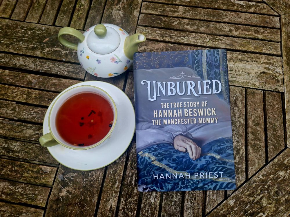 A copy of Unburied: The True Story of Hannah Beswick, the Manchester Mummy by Hannah Priest on a wooden table with a cup of tea and a small teapot next to it.