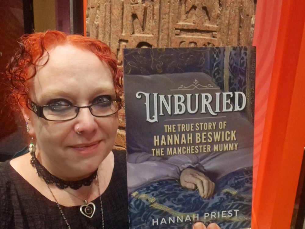 Hannah has red hair, black glasses and a black dress. She is holding a copy of Unburied: The True Story of Hannah Beswick, the Manchester Mummy in front of a pillar in the Manchester Museum.