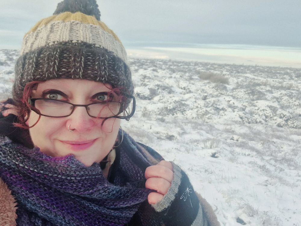 Hannah is wearing a woollen hat, a woollen scarf, fingerless gloves and black glasses. She is standing against a landscape of snow on moorland.
