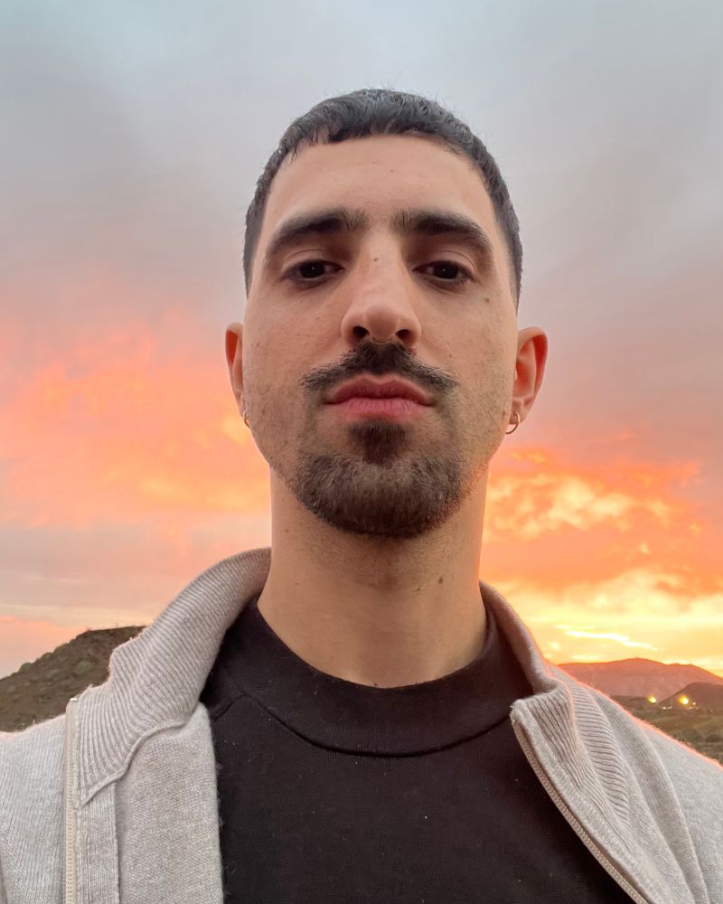 Mediterranean queer boy in the Atlantic Ocean. I’m wearing a beige cardigan and a red shirt, I have a dark moustache and goatee and freshly cut short hair 