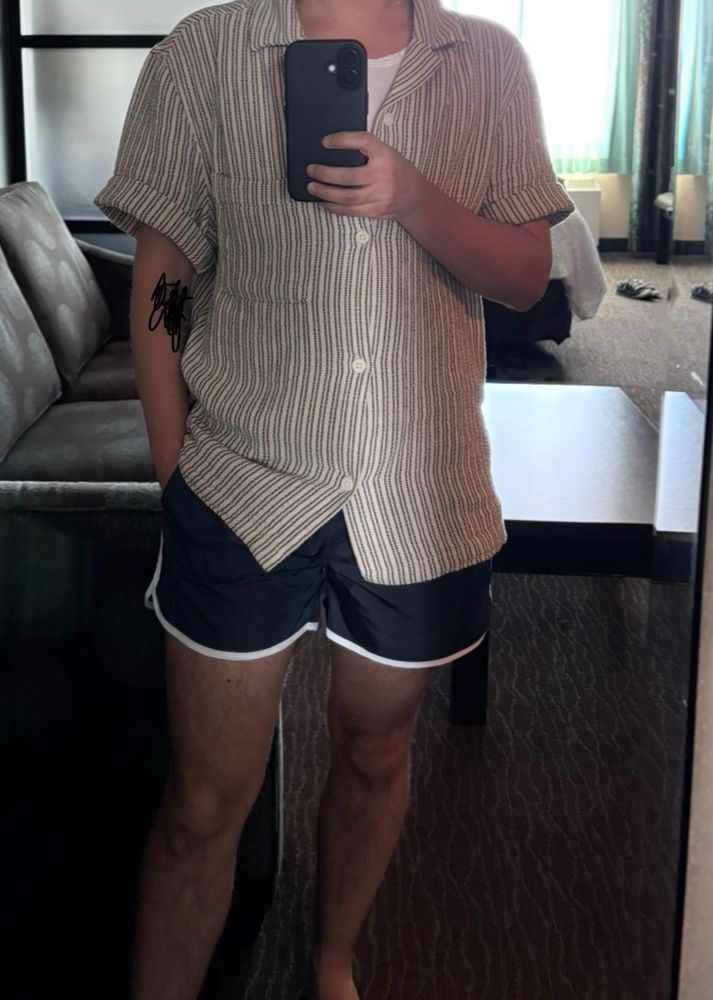 Man taking a photo in a full length mirror. He has navy short shorts on and a knitted short sleeve shirt.