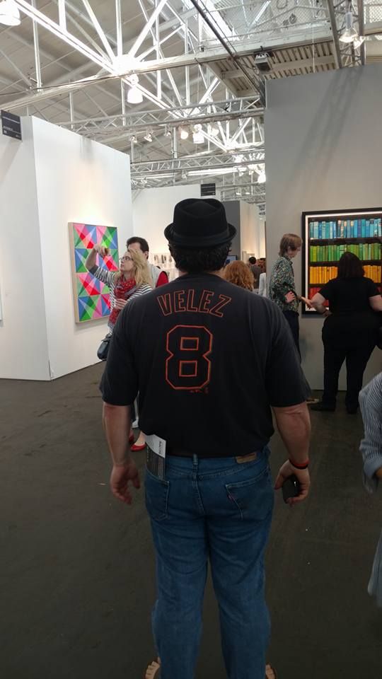 At an art fair, a guy stands wearing a sandals, bluejeans, a mini-fedora, and a black-and-orange Velez #8 Giants shirsey.  

(Cue theme from Succession)