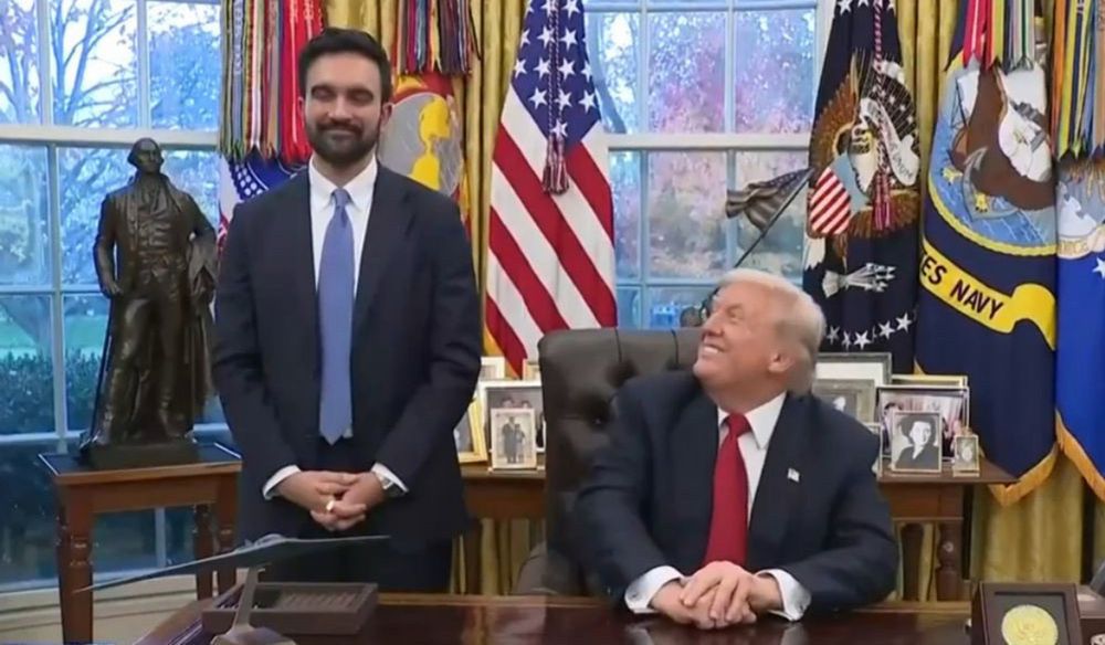A man in a dark suit with a blue tie standing next to another man seated at a desk wearing a dark suit with a red tie.