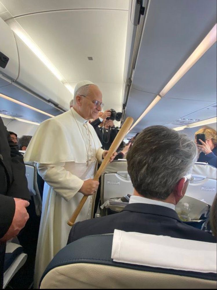 The Pope, standing in the aisle of an airplane, holding a baseball cap.