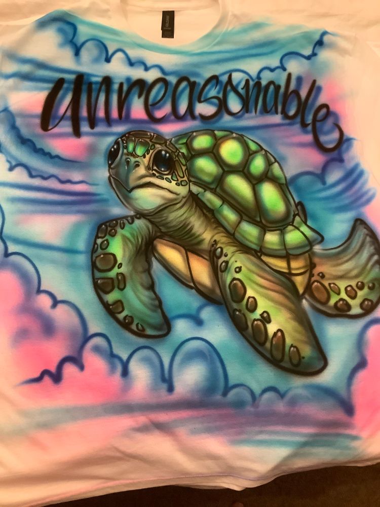 White Tshirt with airbrushed art created by an artist at the fair. flying turtle swimming among pink clouds and blue sky with the word “unreasonable” above