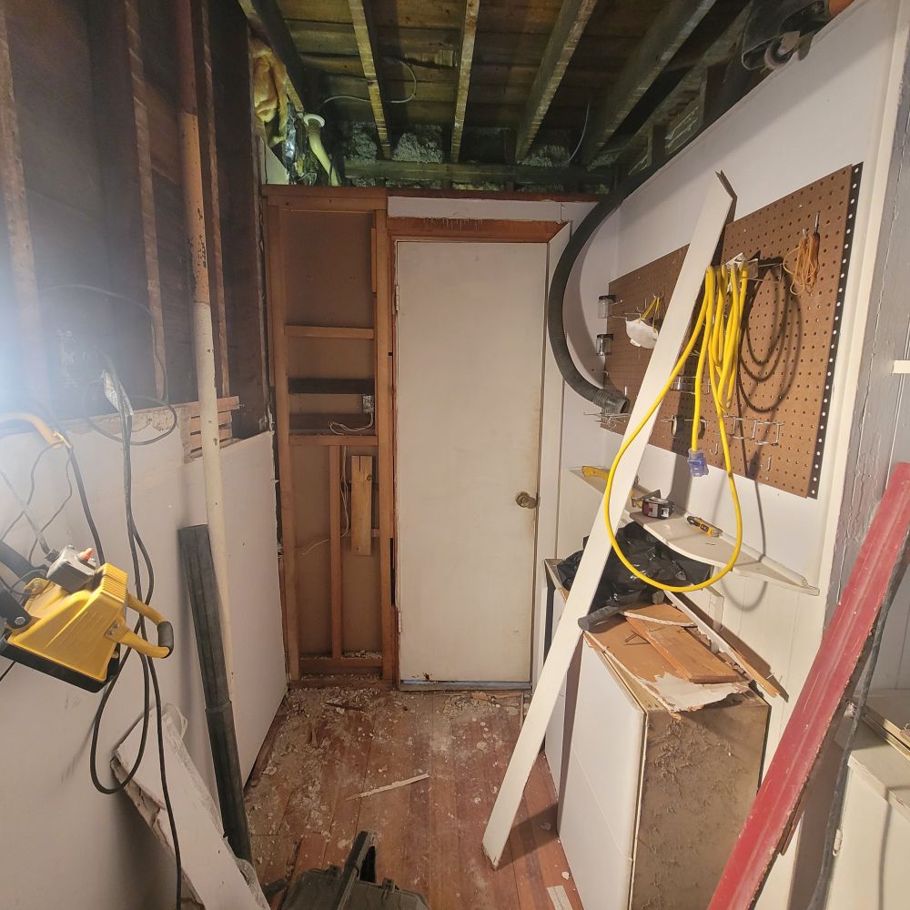 A pantry half way through demolition in a house from the early 1900s, two walls mostly opened up and the ceiling laid bare to the joists. Whites, browns, and a touch of a green glow in the top left corner. 