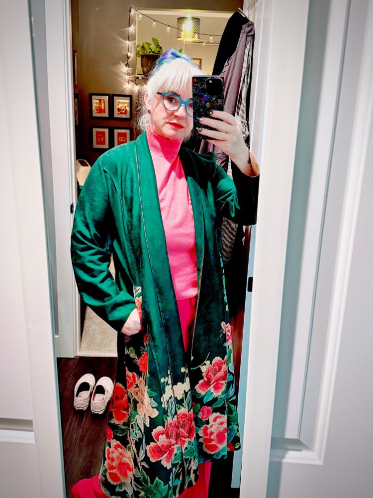 Blonde woman in pink turtleneck and pants, now wearing a green flowered coat and feeling mildly ridiculous. 